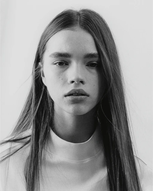 Monochrome portrait of a young woman with long straight hair, fair skin, and a neutral expression, wearing a white mock neck top, against a light background.
