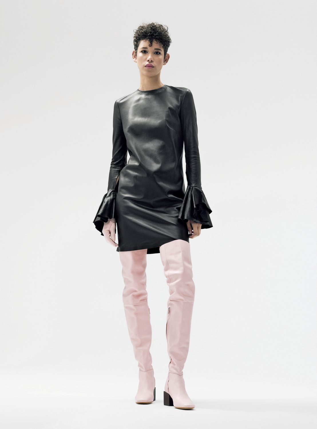 A woman in a black dress and pink thigh-high boots stands in a bright studio setting, showcasing a modern and fashionable aesthetic.