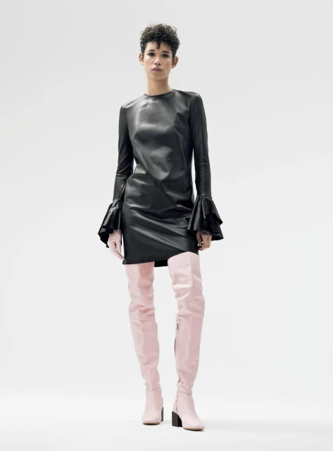 A woman in a black dress and pink thigh-high boots stands in a bright studio setting, showcasing a modern and fashionable aesthetic.