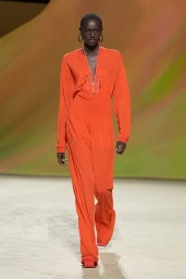 Model walking the runway wearing a vibrant orange jumpsuit and large hoop earrings