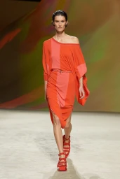 Model walking down a runway wearing an asymmetrical orange dress and red platform sandals with abstract backdrop