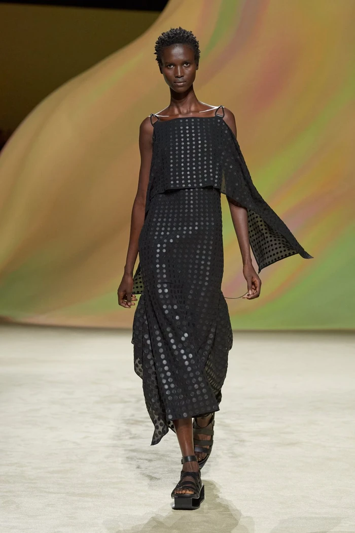 Model on a runway wearing a black patterned dress with draped capelets and strappy sandals