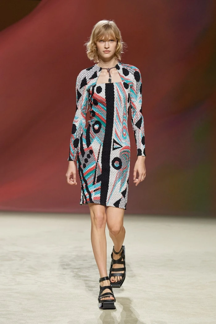 Model walking the runway wearing a long-sleeve patterned dress and black platform sandals on a neutral studio floor.