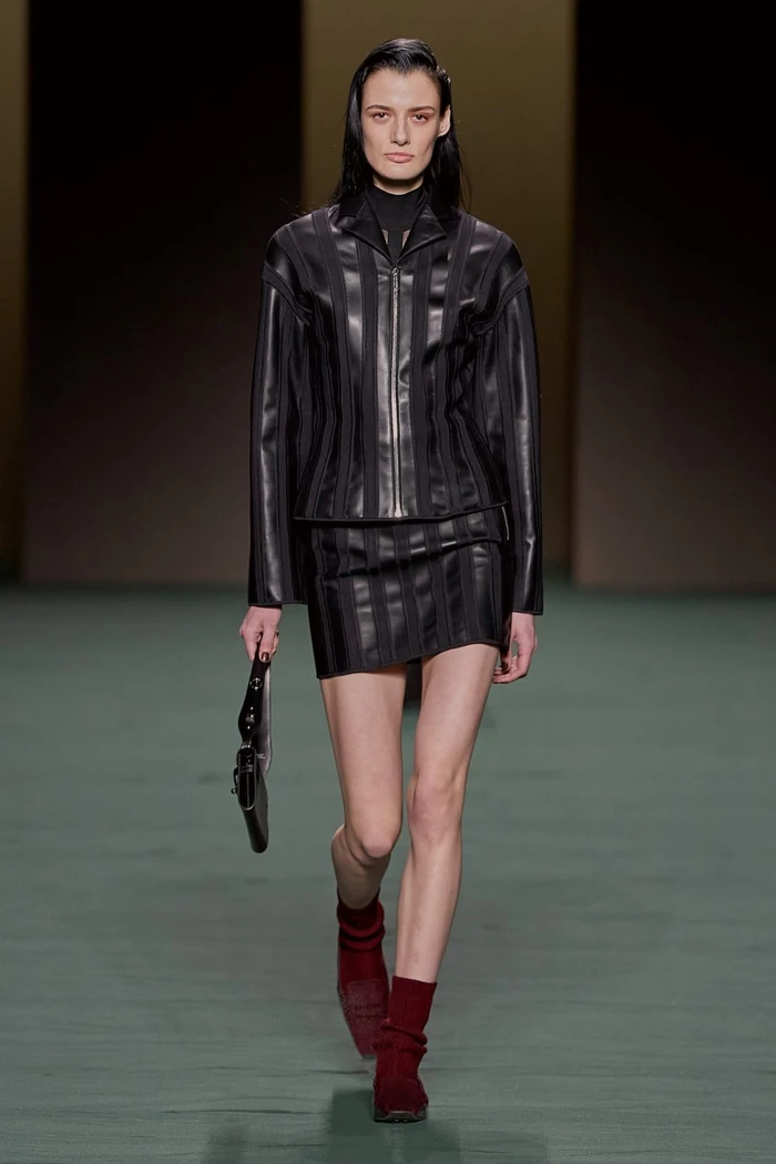 Model in striped black leather jacket and skirt holding a small handbag on runway