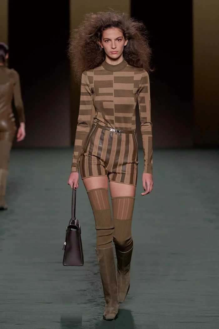 Model walking runway wearing striped brown long sleeve top, matching shorts, knee-high boots, and carrying a handbag