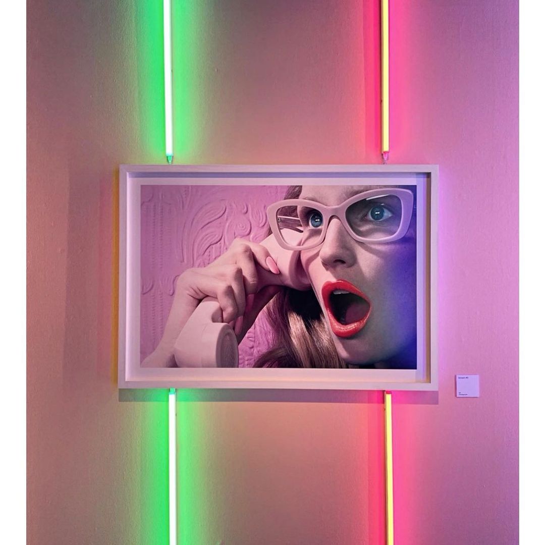 Framed photo of a woman with glasses holding a retro telephone with her mouth open, lit by green and pink neon tubes.