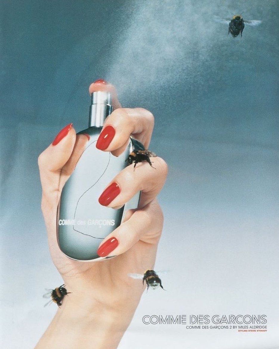 Close-up of a woman's hand holding a perfume bottle, surrounded by bees, set against a soft blue gradient backdrop.