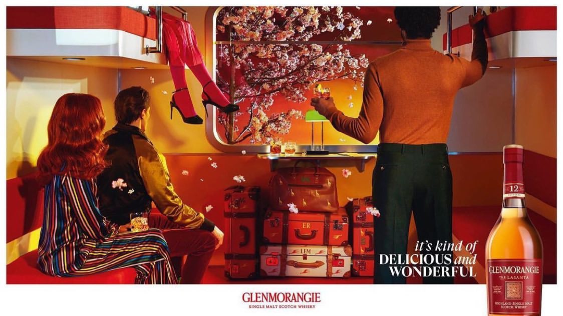 Stylized whisky ad featuring two people seated, another standing, and legs dangling from above in a modern indoor setting with luggage.