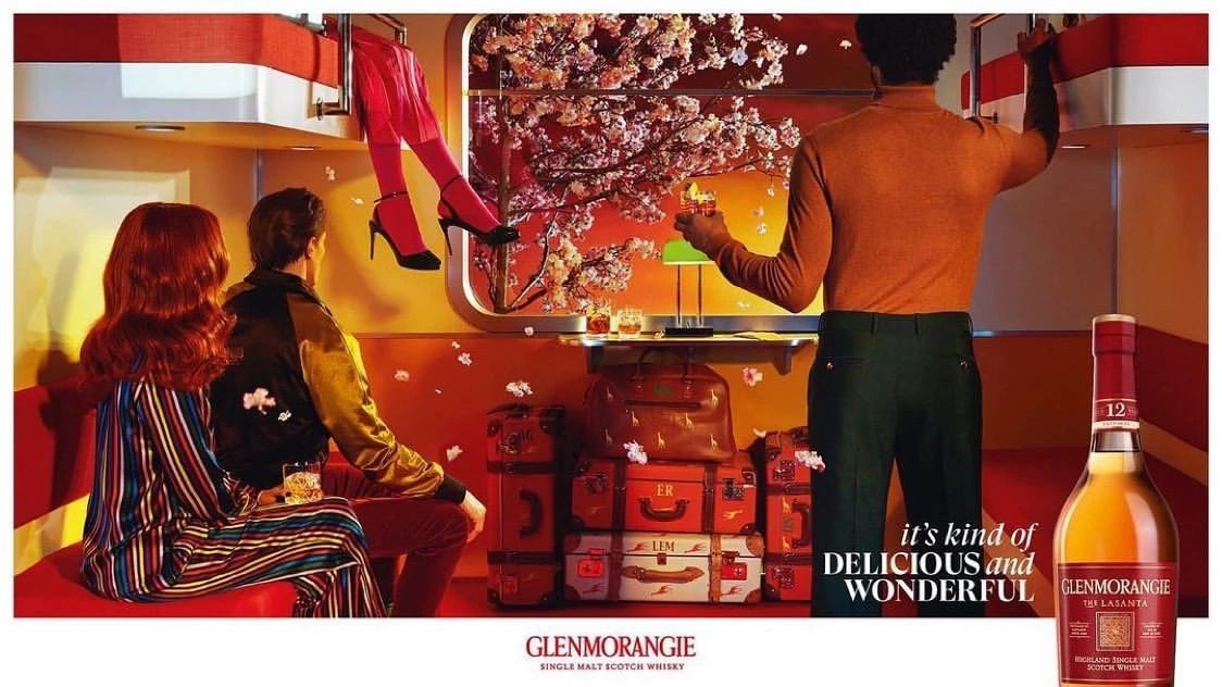 Stylized whisky ad featuring two people seated, another standing, and legs dangling from above in a modern indoor setting with luggage.