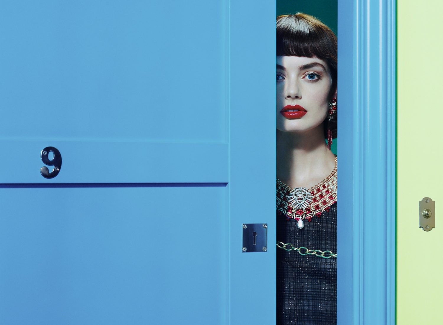 Striking fashion portrait: woman with bold red lipstick and ornate necklace gazes through a vibrant blue doorway, hints of chartreuse.