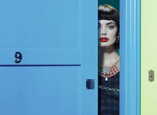 Striking fashion portrait: woman with bold red lipstick and ornate necklace gazes through a vibrant blue doorway, hints of chartreuse.