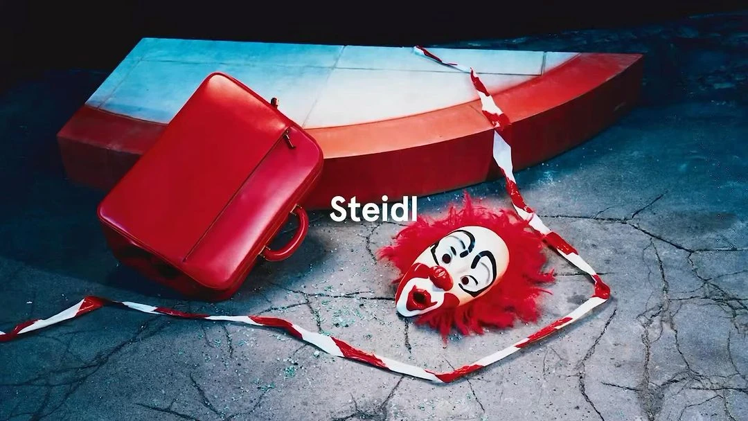 Red briefcase near clown mask with red hair on cracked concrete, in front of a blue-and-red wooden structure, moody lighting.