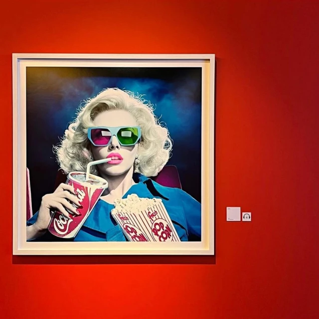 Framed photograph of a woman in 3D glasses drinking soda and holding popcorn, mounted on a red wall in an indoor setting.