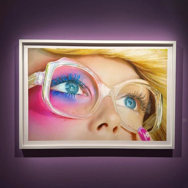 Close-up art print features the upper portion of a woman's face with blond hair, blue eyes, bright eye shadow, and large retro eyeglasses.