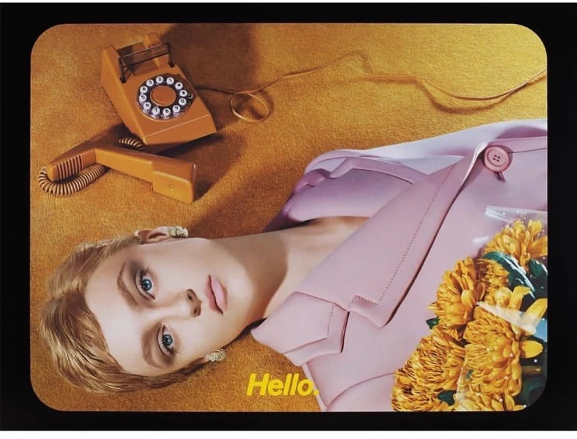Blonde woman in pink blazer lying on yellow carpet next to rotary phone and yellow chrysanthemums in fashion editorial shot.