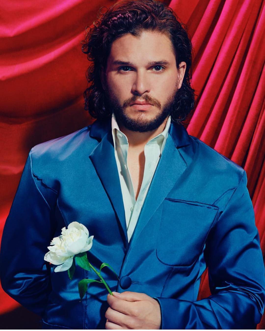 Man in blue suit poses with white flower in a studio setting with red drapes. Portrait-style photography with soft lighting and elegant styling.