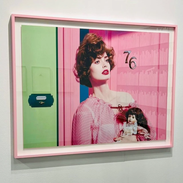 Framed art print featuring a woman in a pink dress holding a doll against a pink and green background, mounted on a white wall.
