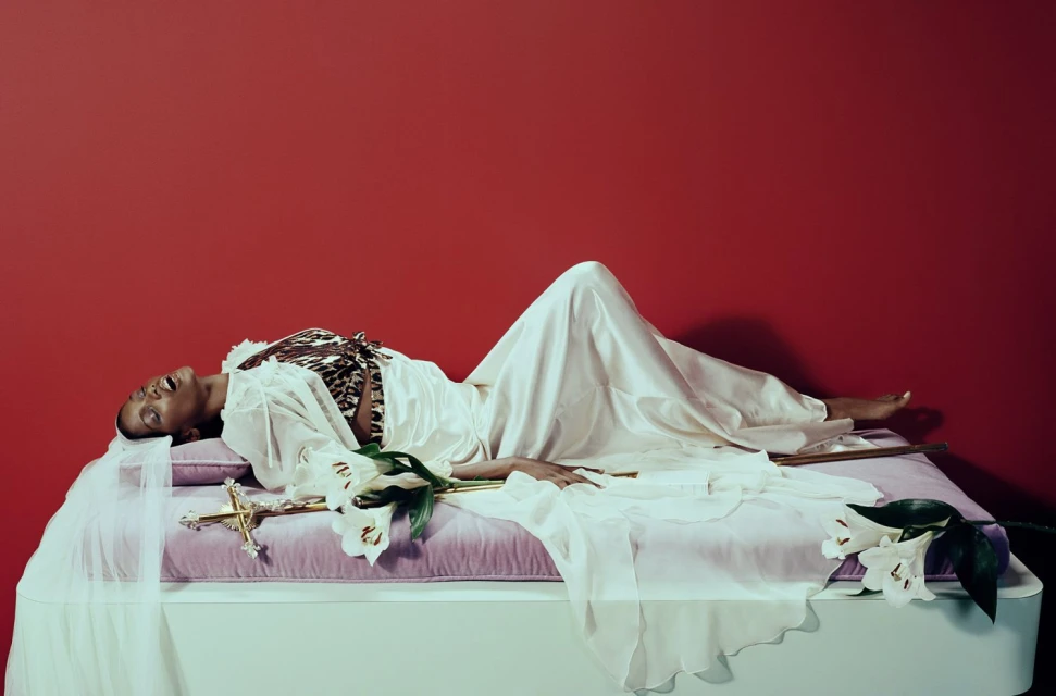 A woman lies on a bed in a white robe with a leopard print top, lilies, and a gold cross against a red wall in a studio setting.