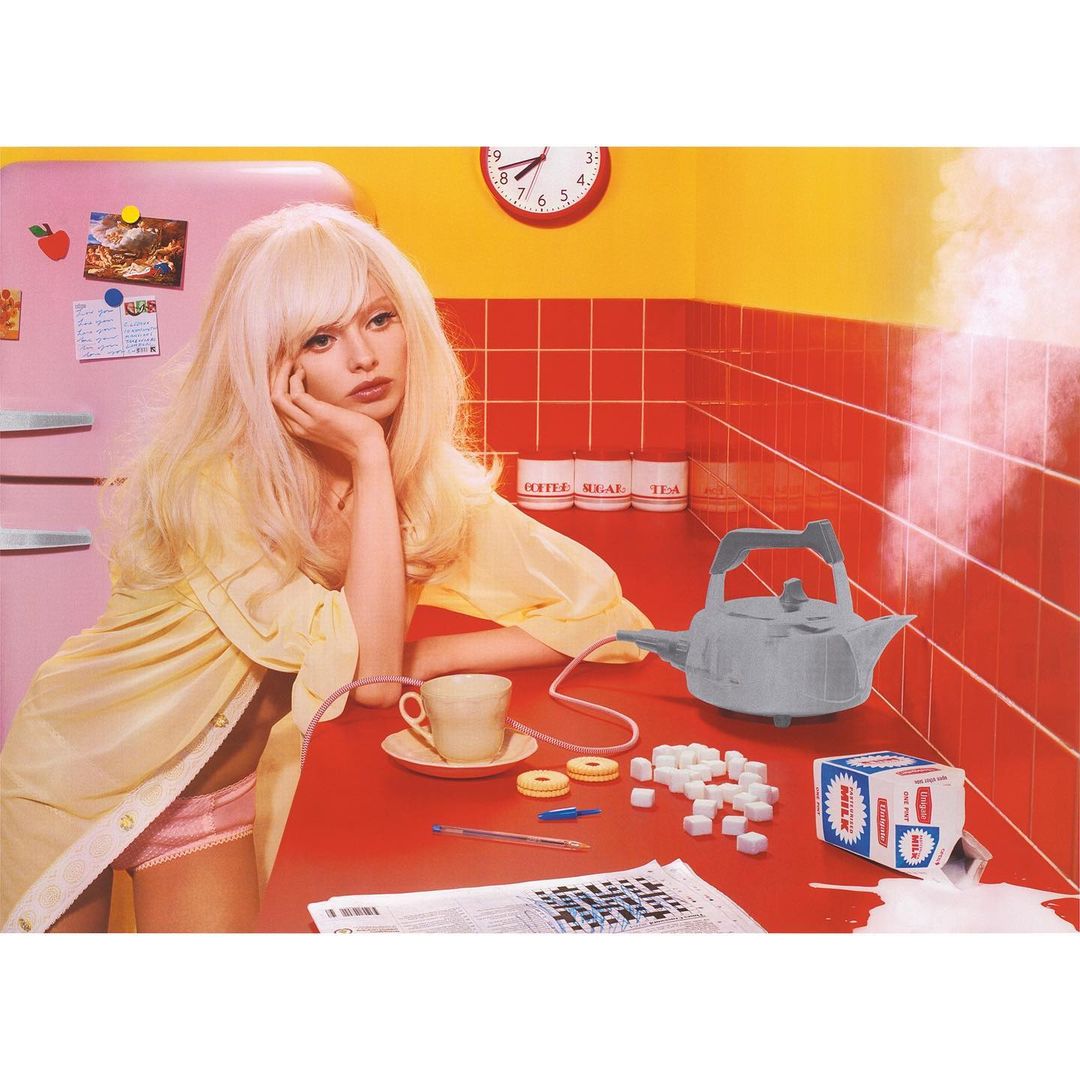 Woman in yellow robe sits at red kitchen counter with tea, sugar cubes, and kettle, creating a retro and colorful, slightly surreal scene.