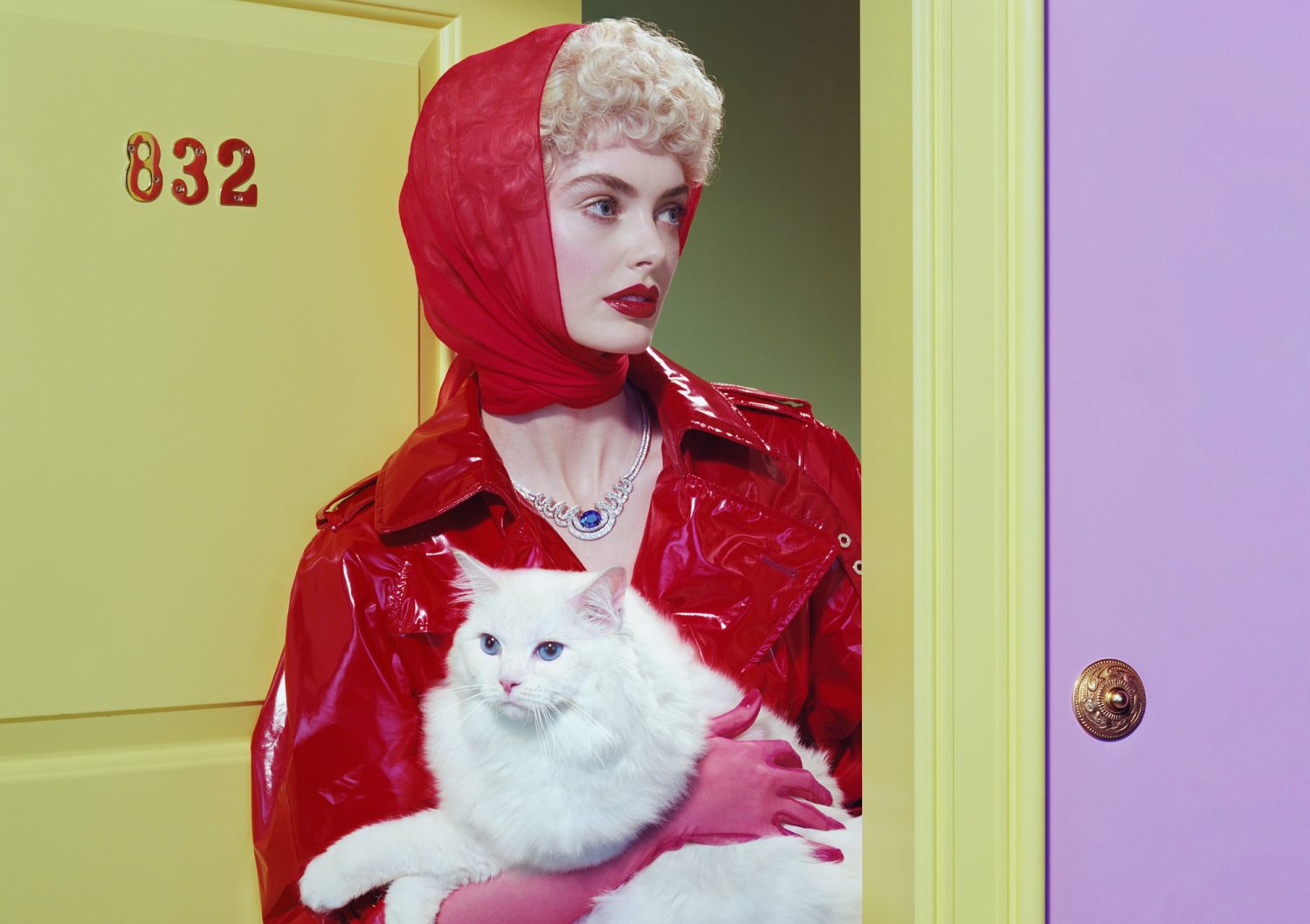 Woman in red coat and headscarf poses with a white cat against split yellow-purple doorway and wall in a studio fashion shot.