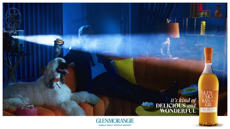 Afghan hound lies on an orange couch next to a man holding a glass, illuminated by a vintage film projector; Glenmorangie Scotch advert.