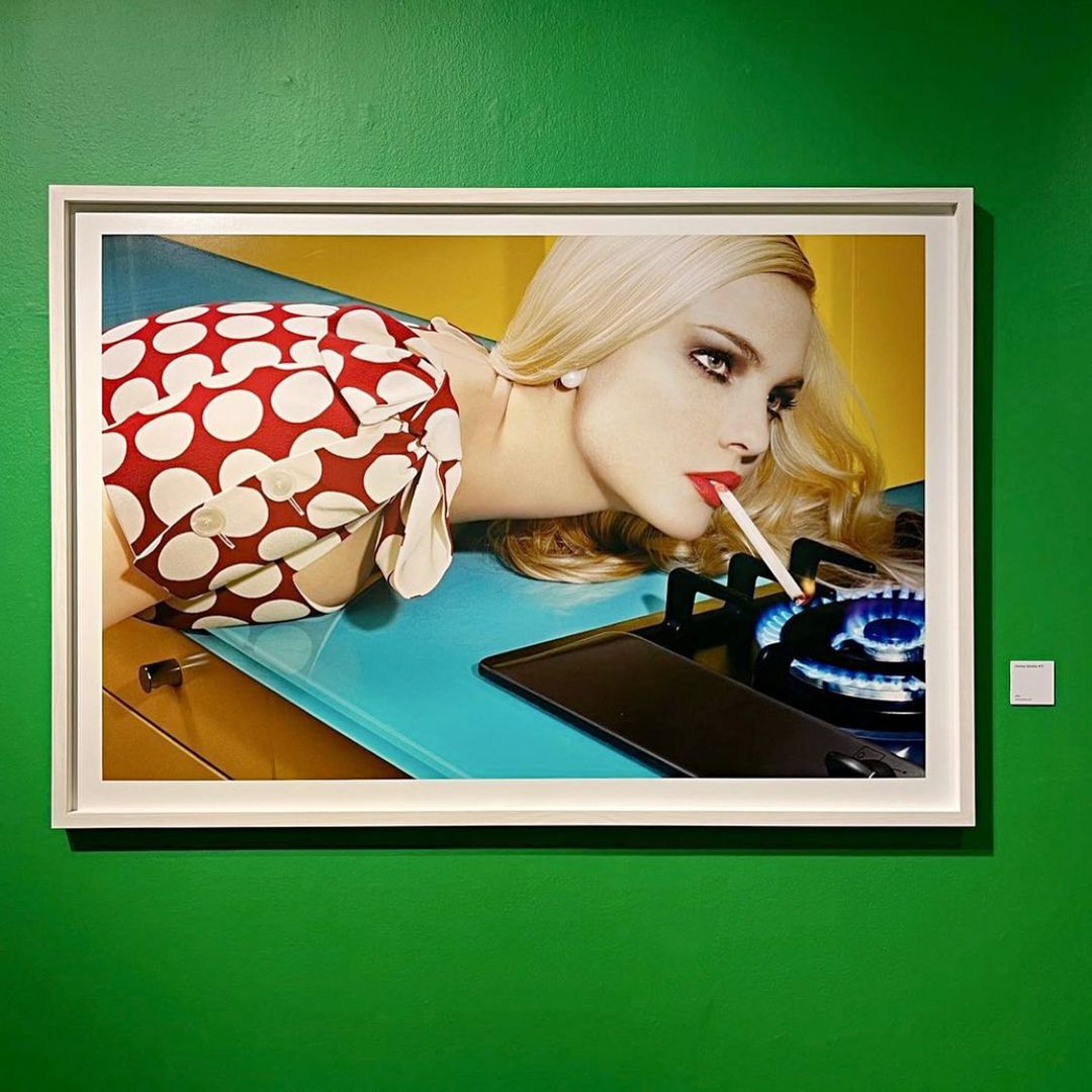 Framed portrait of a blonde woman with red lipstick holding a cigarette, mounted on a green wall in an indoor setting with neutral light.