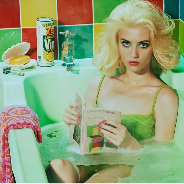 Woman with platinum blonde hair reads a book while relaxing in a vintage-style bathtub, with colorful tiles and retro bathroom accessories.