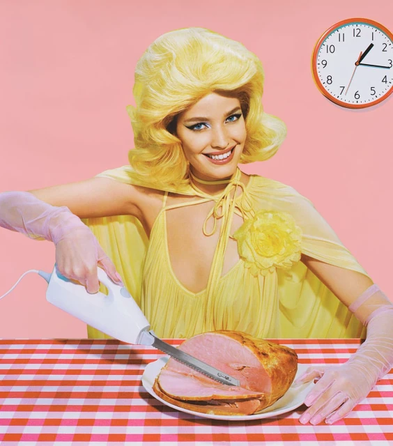 Woman with retro blonde hairstyle slices ham on gingham tablecloth under clock against pink background in stylized, colorful studio shot.