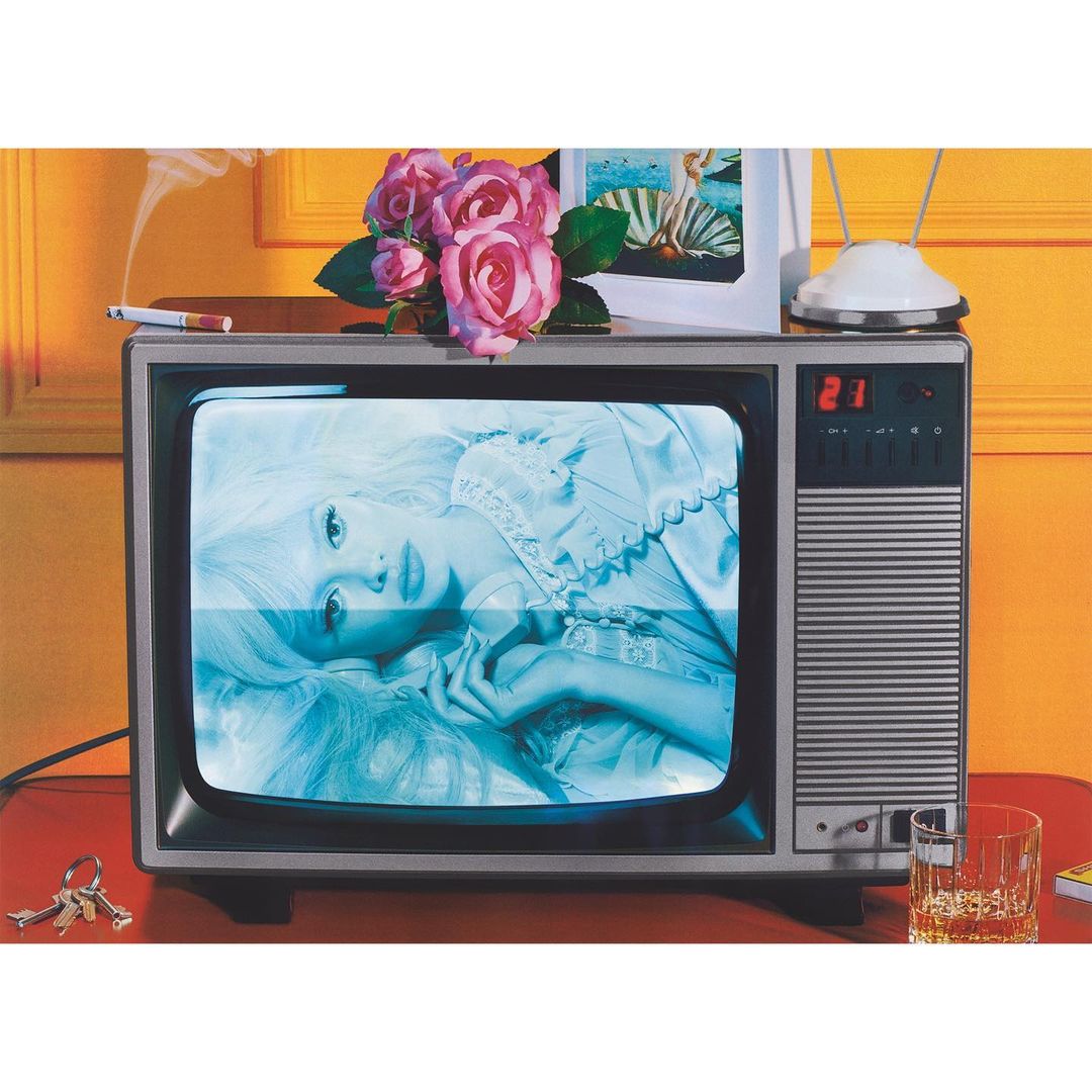 Art photograph of a television set with a woman on the screen, adorned with roses and a cigarette on top, against an orange wall.