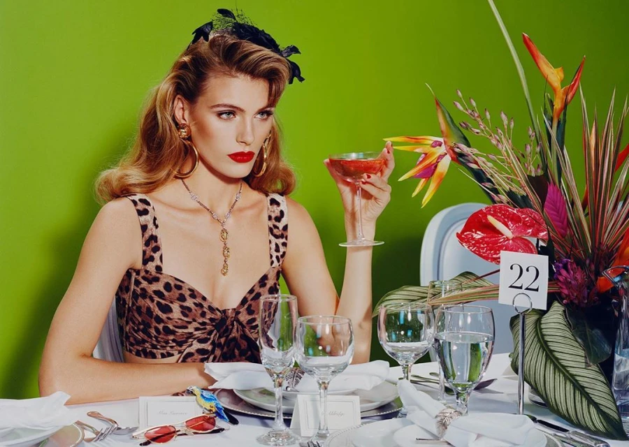 A poised blonde woman with red lipstick in a leopard print top is seated at a formal table, holding a cocktail against a bright green backdrop.