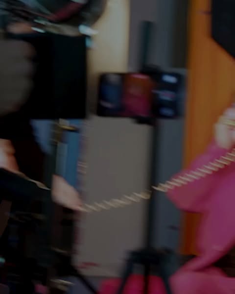 Motion blur captures two people with a cellphone tripod in an indoor environment, one wearing a pink jacket and the other in black.