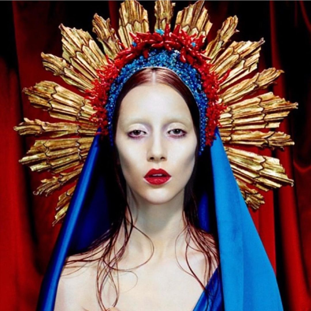 Portrait of a woman with pale skin and red lips wearing an elaborate blue and red crown with a gold ray halo, against red fabric.