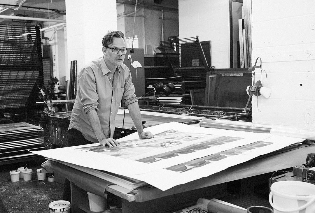Adult man in glasses leaning over a table with large printed sheets, working in a bright workshop with industrial equipment visible.