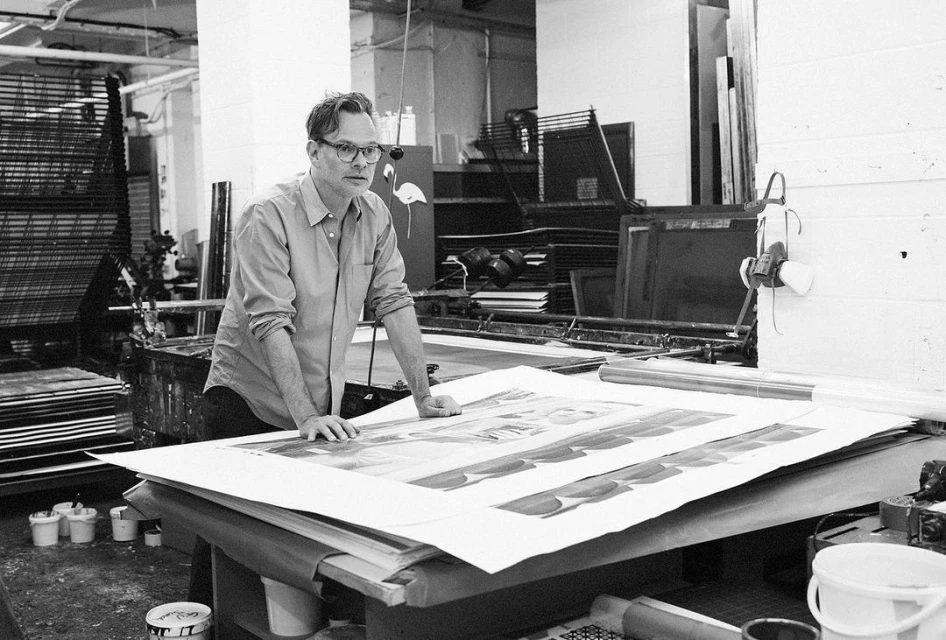 Adult man in glasses leaning over a table with large printed sheets, working in a bright workshop with industrial equipment visible.