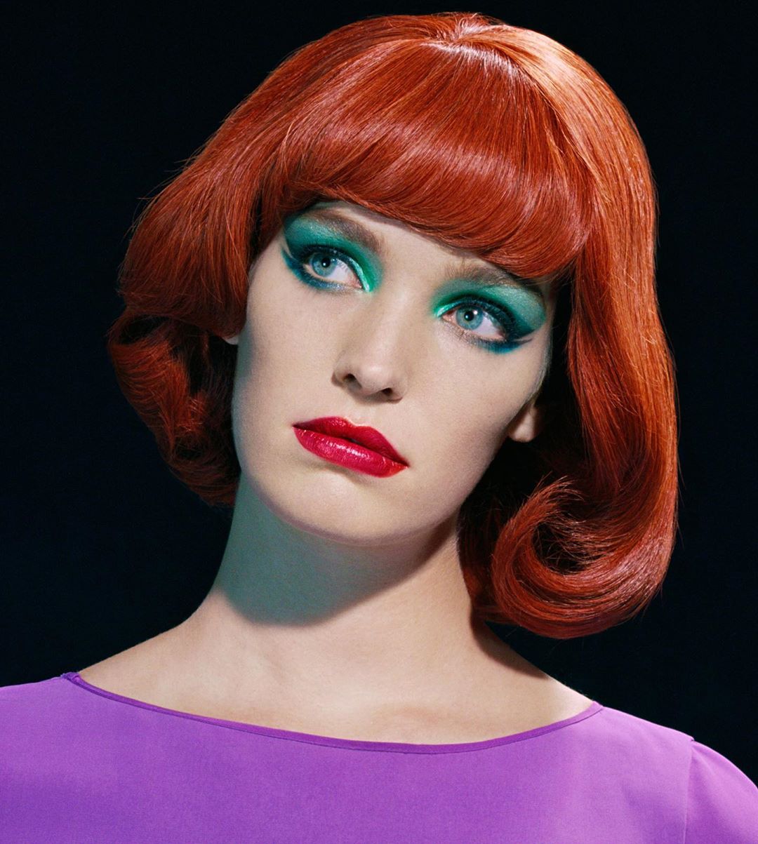 Portrait of a woman with bright red bobbed hair, vibrant teal eyeshadow, and red lipstick wearing a purple top
