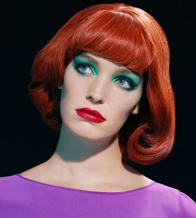 Portrait of a woman with bright red bobbed hair, vibrant teal eyeshadow, and red lipstick wearing a purple top
