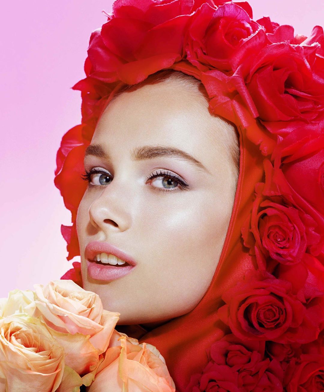 Close-up portrait of a woman with a head covering made of red roses, holding peach-colored roses, against a gradient pink background.