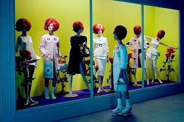 Window display featuring mannequins in vintage-style clothing and wigs, with floral arrangements against a bright yellow backdrop and blue frame.