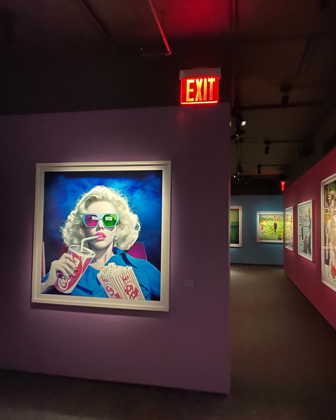 Artwork on a purple gallery wall shows a woman in 3D glasses holding a drink and popcorn with gallery visitors in the background.