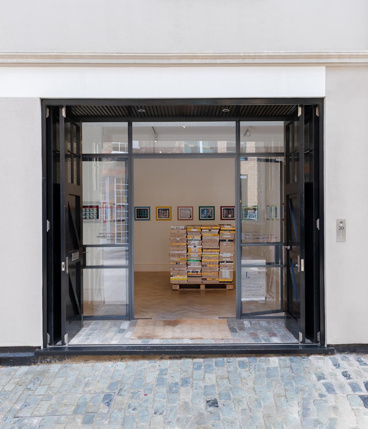Open glass doors leading to an indoor space displaying stacks of product samples and framed images, creating a modern gallery view.