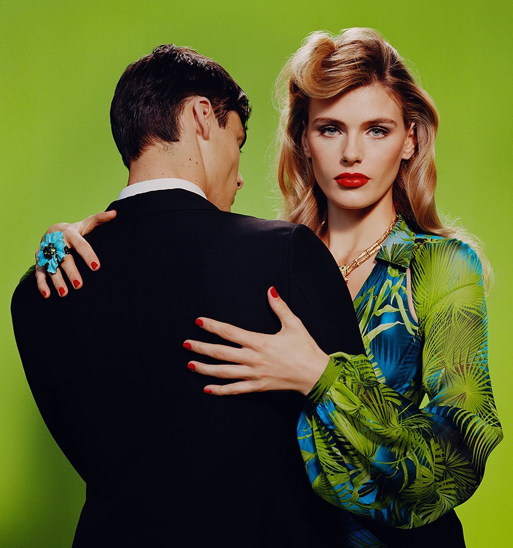 Stylish couple poses in front of a green background. The woman in patterned blouse has red lips and a gold necklace.