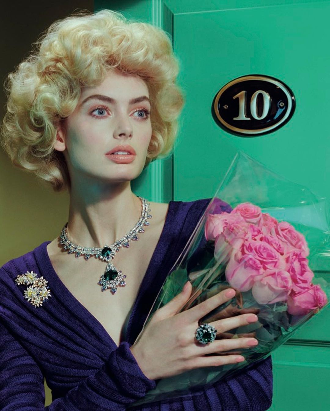 Woman in purple dress holds pink roses near green door with number sign, wearing jewelry: necklace, ring, and flower brooch.