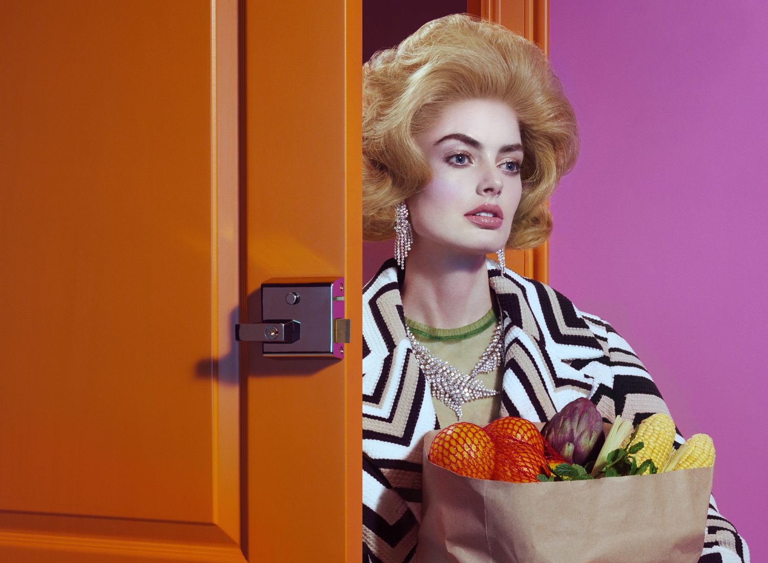 Woman with retro hair and geometric coat holds bag of produce, standing by orange door against a pink wall in a fashion-style photo.