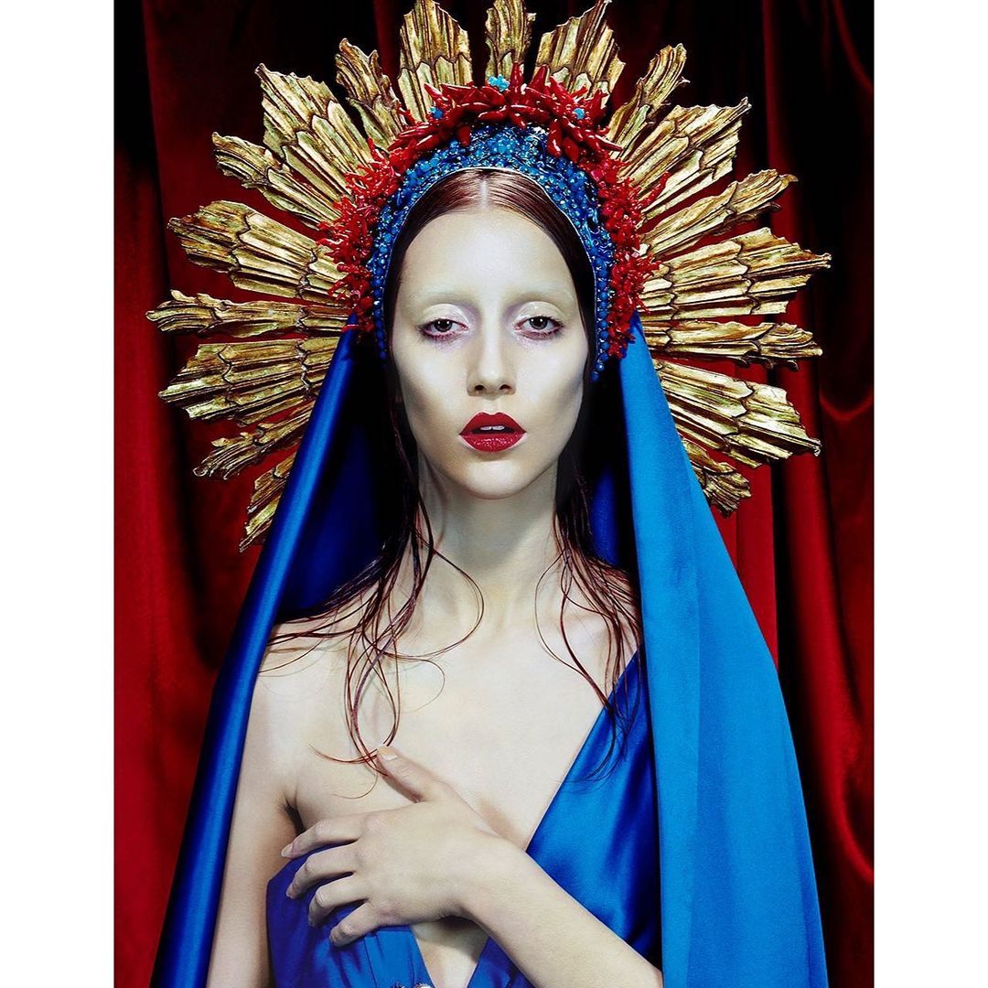 Woman in a blue robe and ornate headpiece in a portrait with a red curtain background. High fashion and beauty shot.