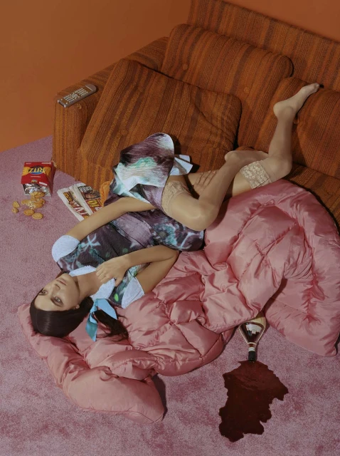 Woman lies on pink duvet on carpet next to spilled wine, crackers, remote and orange couch in a brightly lit indoor scene.