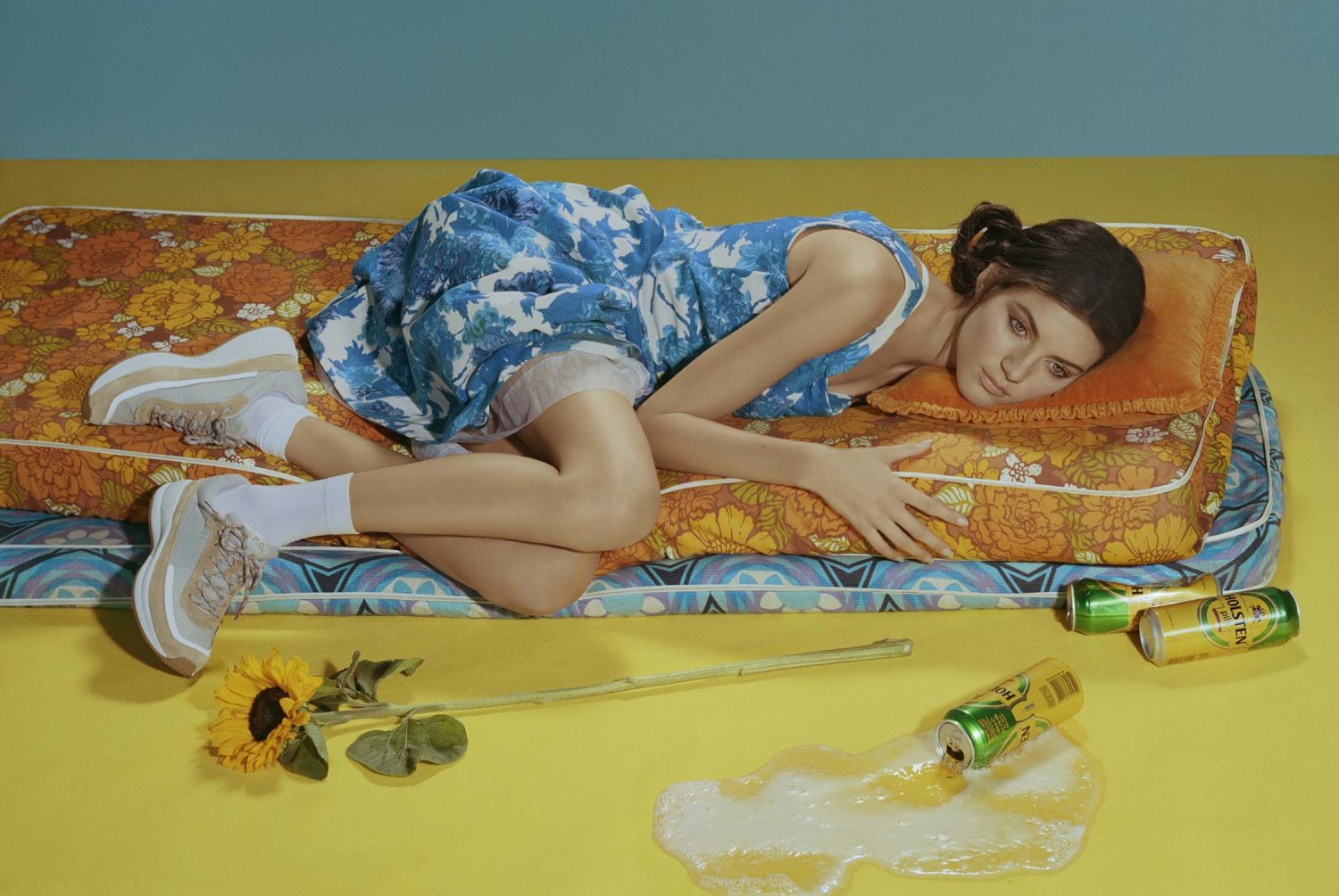 Woman in blue dress lounges on floral mattress with beer cans and sunflower in a yellow and blue indoor studio setting.