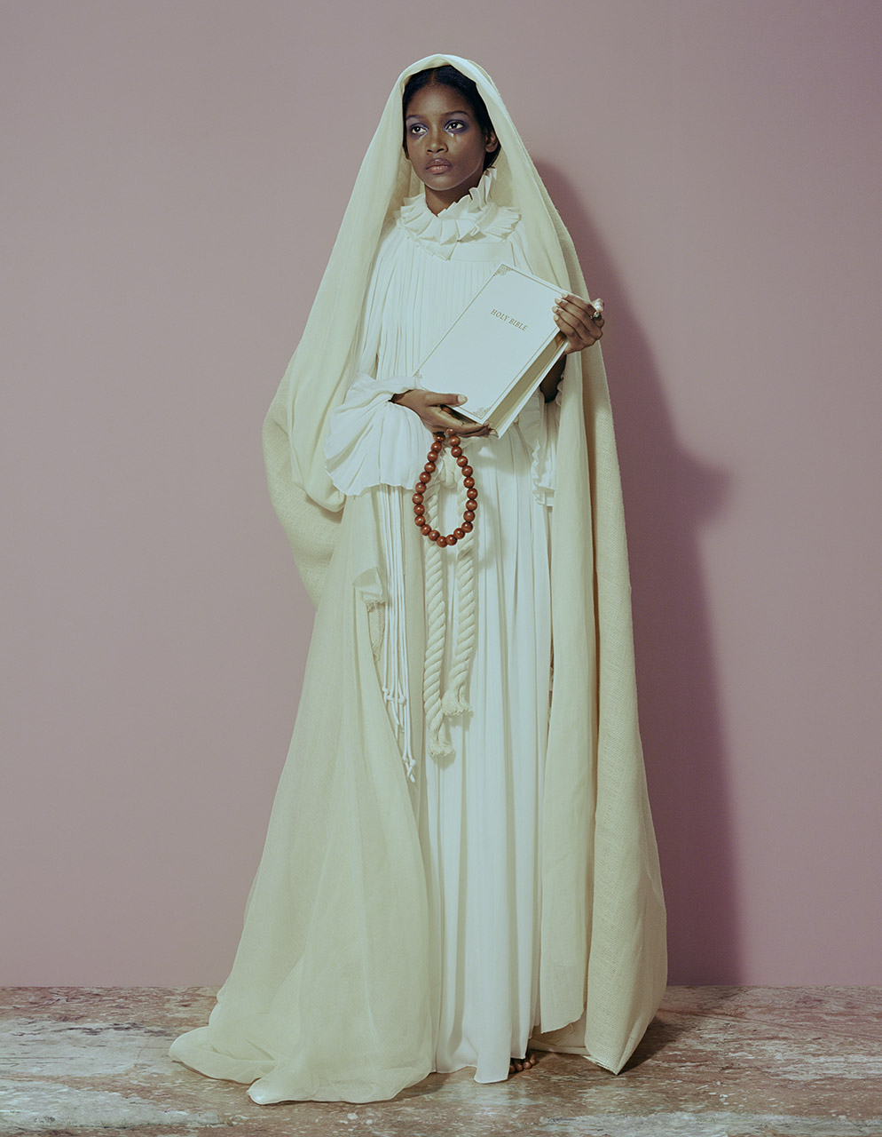 Elegant dark-skinned woman in white gown and headscarf holds a book and wooden bead rope, set against a pink studio backdrop.