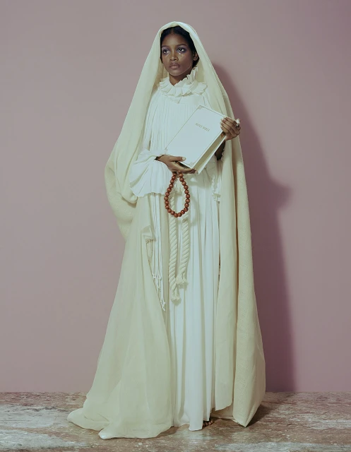 Elegant dark-skinned woman in white gown and headscarf holds a book and wooden bead rope, set against a pink studio backdrop.