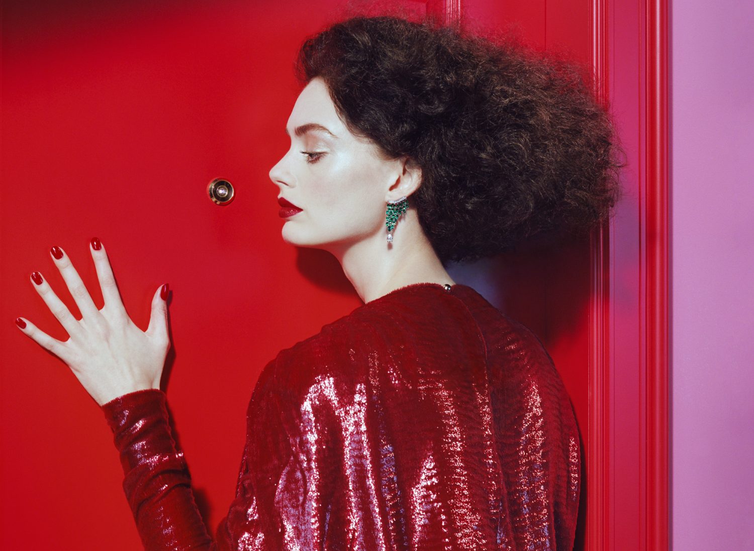 A stylish woman with dark curly hair and bold red lipstick poses in front of a red door, wearing a red sequin dress.