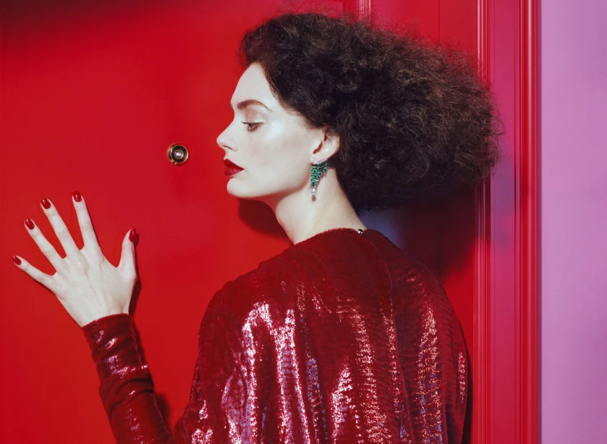 A stylish woman with dark curly hair and bold red lipstick poses in front of a red door, wearing a red sequin dress.